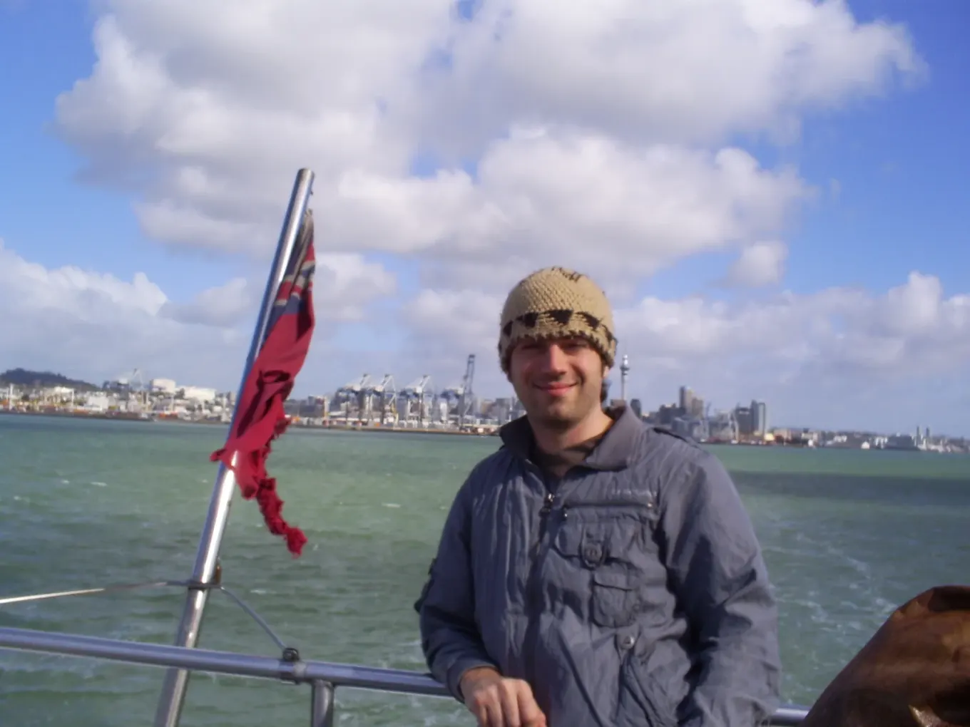 Tom wearing a woolly hat on a boat with Auckland behind him